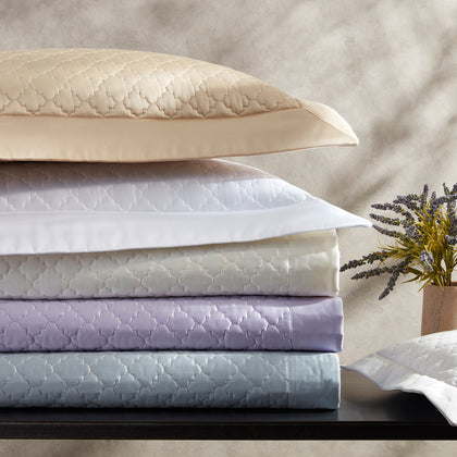 Ava quatrefoil-quilted bedding stacked in gold, white, lavender, and blue, beside a sprig of lavender.