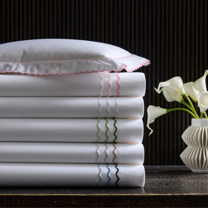 White Axel sheets with wavy embroidered trim in assorted colors, stacked beside calla lilies.
