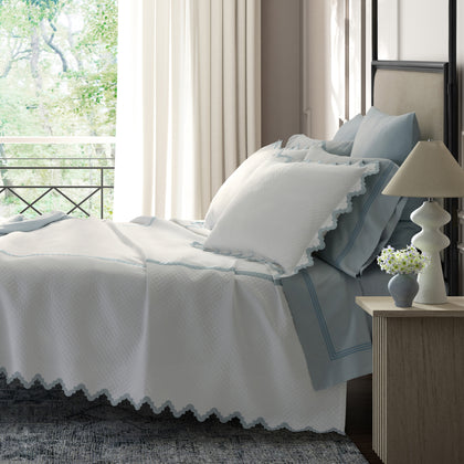 White Aziza Matelasse quilted bedding with blue wavy trim, styled on a bed near a balcony window.