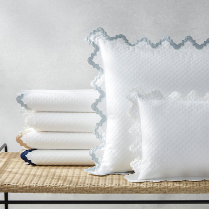 White Aziza Matelasse quilted bedding with gray wavy trim, stacked on a woven bench.