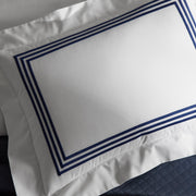 Close-up of a white BelTempo pillow sham with triple navy line embroidered borders, angled against a navy quilted coverlet.