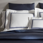 Close-up of stacked white BelTempo pillow shams with triple navy line embroidered borders, against navy quilted pillows and a navy coverlet.