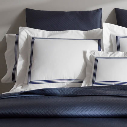 Close-up of stacked white BelTempo pillow shams with triple navy line embroidered borders, against navy quilted pillows and a navy coverlet.