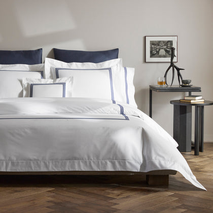 Bed with navy quilted pillows and white BelTempo bedding with triple navy line embroidery, beside a nightstand with a framed photo.
