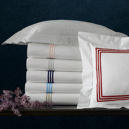 Stack of folded white BelTempo sheets with triple-line embroidery in assorted colors, with a red-trimmed pillow sham and dried flowers.