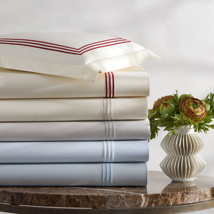 Stacked Bel Tempo Nocturne sheets with striped embroidered trim in ivory and pale blue, beside a vase of flowers.