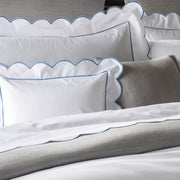 Close-up of white bedding with a scalloped blue embroidered border, gray textured throw pillow.