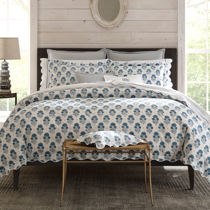 Bed with a blue and white botanical block-print duvet and scalloped edge, striped shams, wood-paneled wall, round mirror, bench with a matching sham.