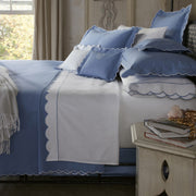 Bed with blue scalloped-edge bedding and a white sheet with a blue scalloped border, monogrammed pillow, nightstand with books and jewelry.