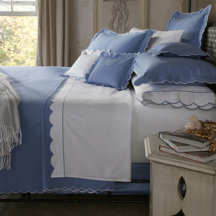 Bed with blue scalloped-edge bedding and a white sheet with a blue scalloped border, monogrammed pillow, nightstand with books and jewelry.