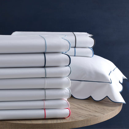 Stacked folded white sheets with thin scalloped borders in light blue, navy, white, and red, beside a navy pillow sham with a scalloped border.