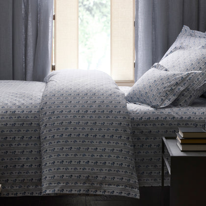 Blue and white Cabanon Stripe bedding styled on a bed beside a curtained window.