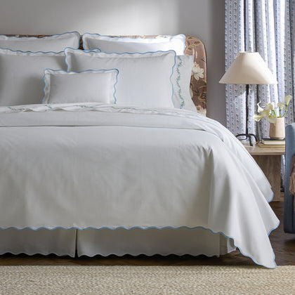White Camilla Pique scalloped bedding with light blue trim, styled on a bed near a floral headboard.