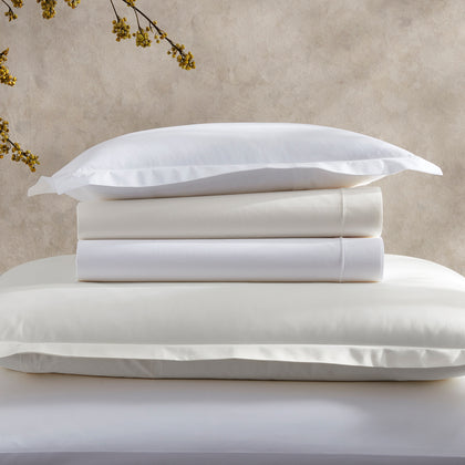 Stacked white and ivory Ceylon percale sheets beside a flowering branch.