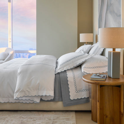 Bed with white bedding bordered in a gray chain-link embroidered scallop, city view window, wood nightstand with a lamp.