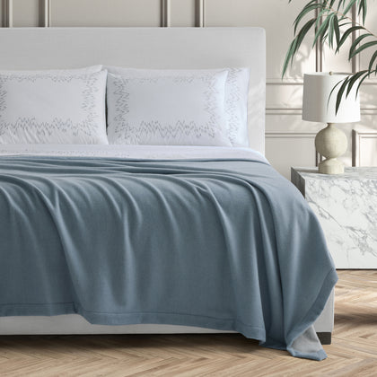 Slate blue Cosmo blanket draped on a bed with zigzag-embroidered shams.
