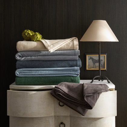 Cosmo blankets folded in tan, gray, navy, and green, stacked on a chest beside a horse painting.