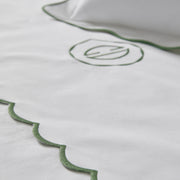 Close-up of white bedding with a scalloped green border and a stylized circular monogram.