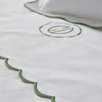 Close-up of white bedding with a scalloped green border and a stylized circular monogram.