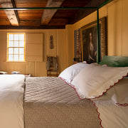 White bedding with a scalloped red edge, taupe quilted coverlet, green four-poster bed, rustic room with a wood beam ceiling, antique portrait.
