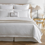 Bed with white bedding and a tan scalloped edge, gray Greek-key bolster pillow with a "PMO" monogram, tan upholstered headboard.