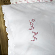 White pillow sham with a scalloped edge, embroidered "Daisy May" in pink script.