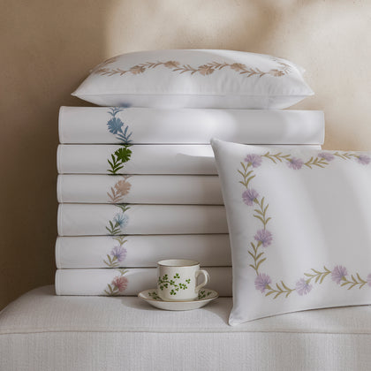 White Daphne sheets stacked with embroidered floral trim in assorted colors, beside a teacup.