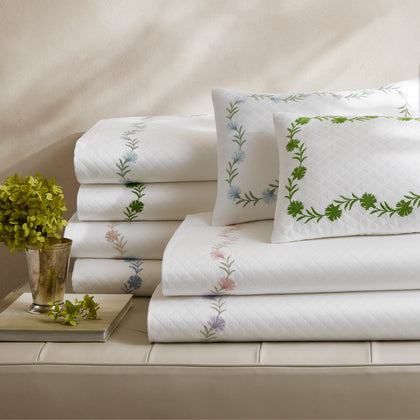 White Daphne Matelasse quilted sheets with floral embroidery, stacked on a bench beside a plant.