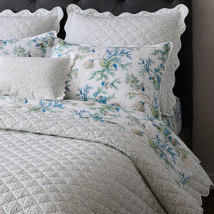 Blue Del Tesoro coral and shell print pillows with scalloped speckled shams, styled on a bed with a dark tufted headboard.