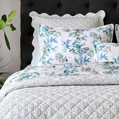 Close-up of blue Del Tesoro coral and shell print pillows with a speckled quilted sham, against a dark tufted headboard.