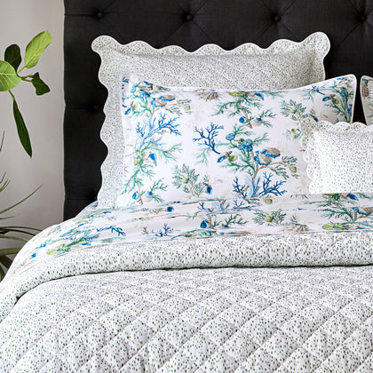 Blue Del Tesoro coral and shell print bedding with a speckled quilted coverlet, styled on a bed with a dark tufted headboard.