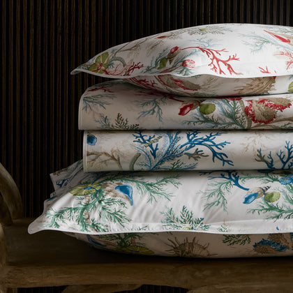 Del Tesoro coral and shell print sheets in red and blue, stacked on a wooden bench.