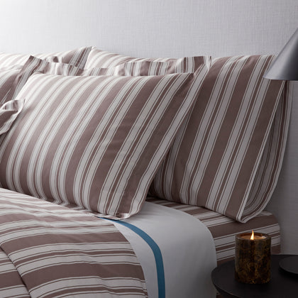 Taupe and white striped pillow shams on a bed with a matching duvet and a white sheet with a blue stripe, lit candle on a side table.