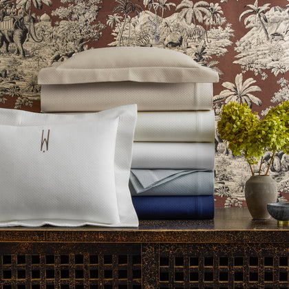Elliot textured pique sheets in tan, white, gray, and navy, stacked beside a monogrammed pillow.