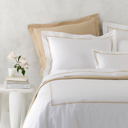 Bed dressed in white Essex bedding with a gold double-line embroidered border and a tan textured throw, beside books and peonies.