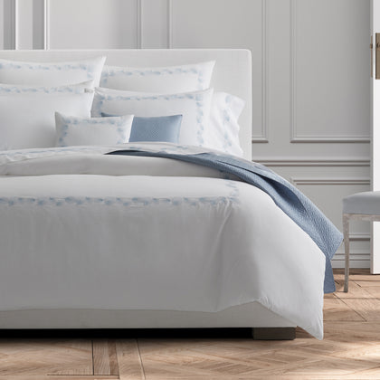 White Feather bedding with blue feather-embroidered trim, styled on a bed with a blue throw.