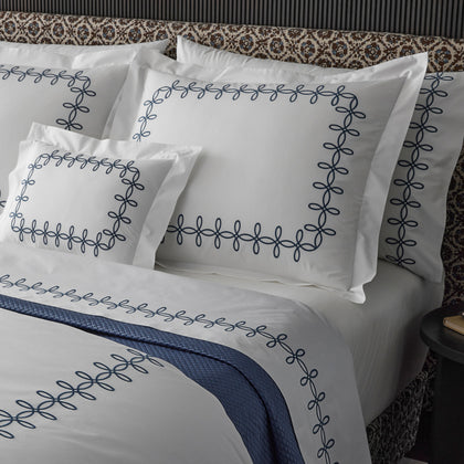 Close-up of white GordianKnot bedding with a navy interlocking-loop embroidered border, styled on a bed with a patterned brown-and-white headboard.