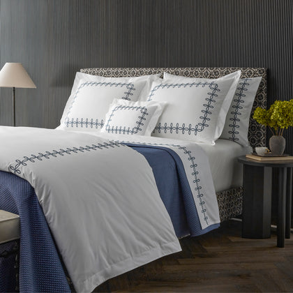 White Gordian Knot bedding with loop-embroidered border, styled on a bed with a navy throw.