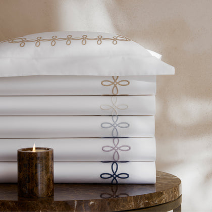 Stack of folded white GordianKnot sheets with interlocking-loop embroidered borders in tan, gray, blue, and navy, with a lit candle on a marble table.