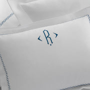 Close-up of a white Hatch boudoir pillow with a navy monogram and diamond-stitch embroidered border, beside stacked pillows.