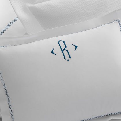 Close-up of a white Hatch boudoir pillow with a navy monogram and diamond-stitch embroidered border, beside stacked pillows.