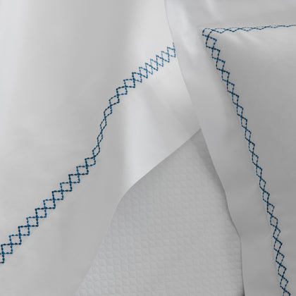 Close-up of white Hatch sheets and pillowcases with blue zigzag embroidered borders, draped over a textured white blanket.