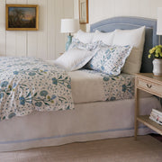 Bed dressed in a blue floral Khilana duvet and quilted white shams, on a blue upholstered headboard, by a nightstand with flowers.