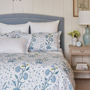 Bed dressed in a blue floral Khilana duvet and shams, on a blue upholstered headboard, by a nightstand with a clock and flowers.