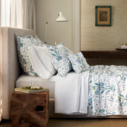 Bed with blue floral Khilana shams and a white sheet with a gray embroidered border, on a tan headboard by a wood stool with a book.