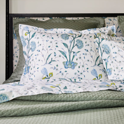 Blue floral Khilana shams layered over a sage green quilted coverlet, on a black iron headboard.