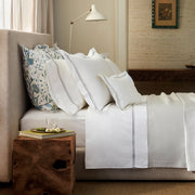 Tan upholstered bed with white sheets bordered in gray double lines and a quilted coverlet, one blue floral Khilana sham at the side.