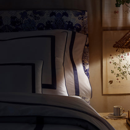White Louise bedding with navy ribbon trim, styled on a bed with a blue-and-white patterned headboard, with botanical prints on the wall.