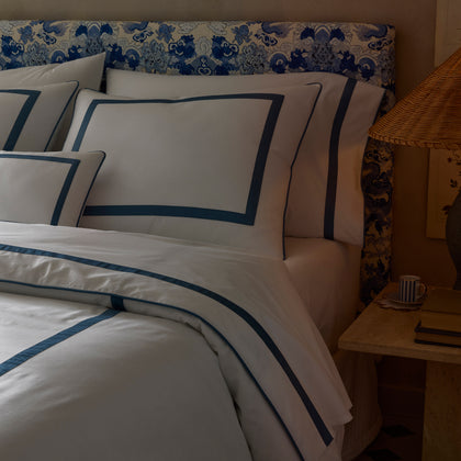 White Louise bedding with blue ribbon trim, styled on a bed with a blue-and-white patterned headboard and a stone nightstand.