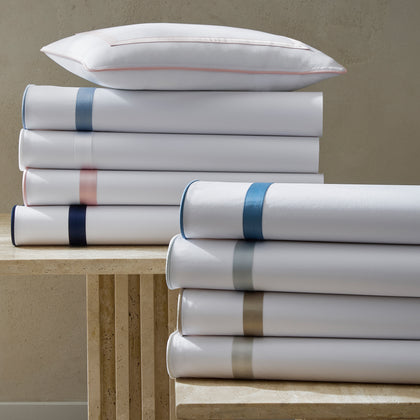 White Louise sheets stacked with satin trim in assorted colors, topped with a pink-bordered pillow.